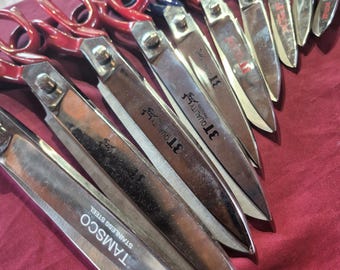 Könnte beinhalten: Eine Sammlung von Edelstahlscheren mit roten und blauen Griffen, die in einer Reihe angeordnet sind. Die Klingen tragen eingravierte Texte wie TAMSCO STAINLESS STEEL und 3T QUALITY. Die Scheren variieren in der Größe und sind diagonal angeordnet.