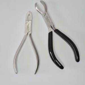 May include: Two stainless steel pliers with curved jaws. One pair has black handles. The text "STAINLESS STEEL PAKISTAN" is printed on the metal. These tools are likely used for jewelry making or other crafts.