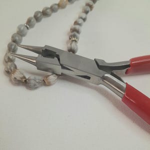 Op de afbeelding: Close-up van een sieradentang met rode handgrepen, geplaatst in de buurt van een kralenketting. De tang is zilverkleurig en lijkt in gebruik te zijn, mogelijk voor het maken of repareren van sieraden. De ketting heeft grijze en witte kralen.