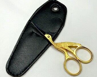 May include: Gold-coloured, bird-shaped, small scissors with a black leather carrying case. The scissors have a detailed design and are closed.