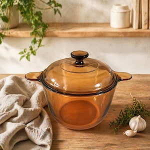 May include: Amber-colored glass casserole dish with a matching lid. The dish has two handles and a dark brown knob on the lid. A beige linen cloth is draped on the wooden surface next to the dish. The dish is designed for cooking.