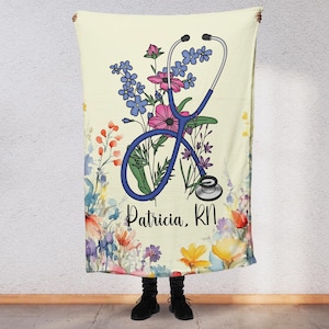 Puede incluir: Una manta de color amarillo claro con un estetoscopio azul y flores de colores. El texto "Patricia, RN" está impreso en la manta.