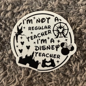 May include: A circular white sticker with black text that reads I'M NOT A REGULAR TEACHER I'M A DISNEY TEACHER. The design includes black silhouettes of a fairy, a Ferris wheel, a castle, a mouse-eared head, and hand gestures forming a heart.