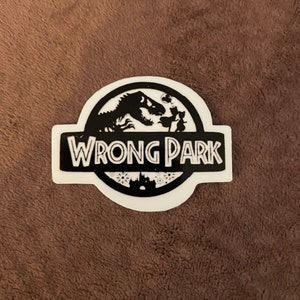 May include: A white sticker with a black graphic of a dinosaur and the words "WRONG PARK" in a stylized font. The sticker also features silhouettes of people and a castle.