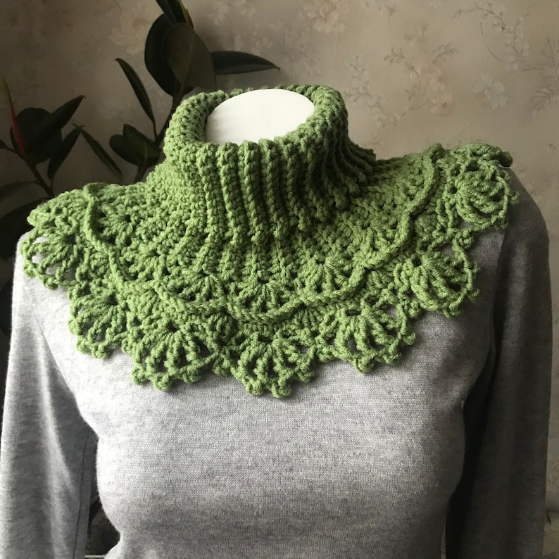 May include: A handmade green crochet cowl with a high, ribbed turtleneck and a decorative scalloped edge. The cowl is displayed on a mannequin and is made of soft, textured yarn. The color is a vibrant, spring-like green.