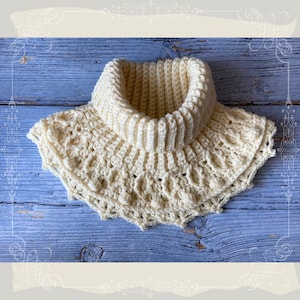 Puede incluir: Un cuello de punto de ganchillo de color crema con un borde rizado y un cuello alto. El cuello está hecho con un punto texturizado y tiene pequeñas bolitas blancas alrededor del borde.