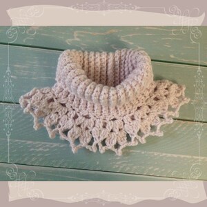 May include: A crocheted cowl in a pale pink colour with a delicate lace pattern around the bottom edge. The cowl is photographed on a light blue wooden surface.