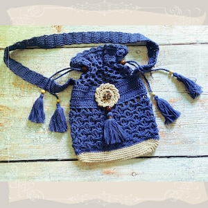May include: A handmade blue crochet bag with a drawstring closure and a long strap. The bag features a beige bottom trim, a beige flower applique, and blue tassels. The bag is made of a soft, textured yarn.