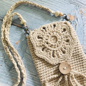 May include: A beige crochet crossbody bag with a decorative floral design on the front flap. The bag features a wooden button closure and a braided strap with wooden bead accents. The bag is designed to hold a phone.