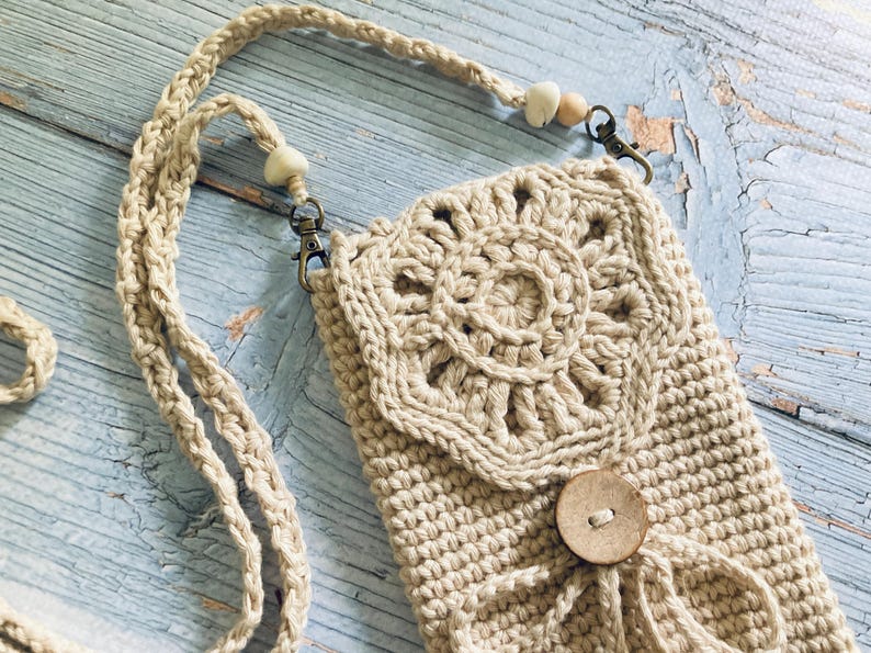May include: A beige crochet crossbody bag with a decorative floral design on the front flap. The bag features a wooden button closure and a braided strap with wooden bead accents. The bag is designed to hold a phone.