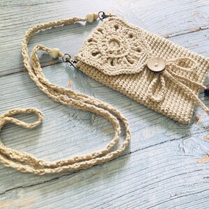 May include: A beige crochet crossbody bag with a floral design on the flap. The bag features a wooden button closure, a braided strap, and decorative tassels with wooden beads. The bag is made of a soft, natural fiber.
