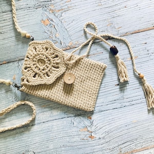 May include: A beige crochet crossbody bag with a floral design on the flap. The bag features a wooden button closure, a woven strap, and decorative tassels. The bag is made of a natural fiber and has a bohemian style.