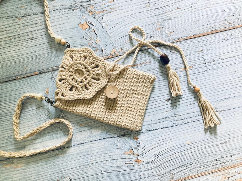 May include: A beige crochet crossbody bag with a floral design on the flap. The bag features a wooden button closure, a woven strap, and decorative tassels. The bag is made of a natural fiber and has a bohemian style.