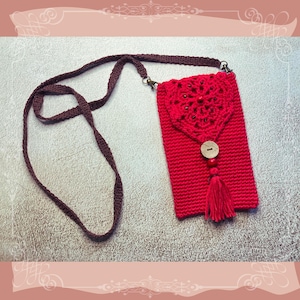 May include: A red crochet phone bag with a brown strap. The bag has a decorative flap with a heart-shaped design, a wooden button closure, and a tassel. The strap is adjustable. The bag is suitable for carrying a mobile phone.