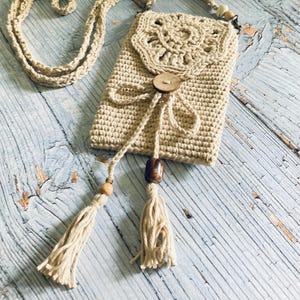 May include: Beige crochet phone bag with a long strap. The bag features a decorative flap with a wooden button closure and tassel accents. The bag is made of a soft, textured yarn and is designed to be worn crossbody.