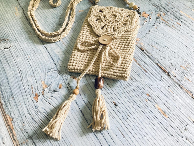 May include: Beige crochet phone bag with a long strap. The bag features a decorative flap with a wooden button closure and tassel accents. The bag is made of a soft, textured yarn and is designed to be worn crossbody.