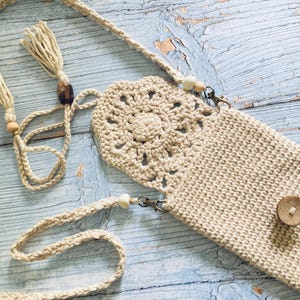 May include: A beige crochet crossbody phone bag with a decorative floral design on the front flap. The bag has a long, braided strap with tassels and wooden beads. A small wooden button secures the bag's closure.