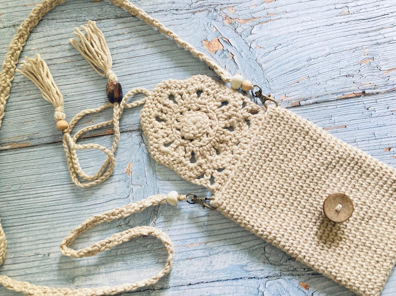 May include: A beige crochet crossbody phone bag with a decorative floral design on the front flap. The bag has a long, braided strap with tassels and wooden beads. A small wooden button secures the bag's closure.