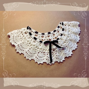 Op de afbeelding: Een crèmekleurige, gehaakte kraag met een geschulpte rand. De kraag heeft een delicaat bloemmotief en is versierd met zwarte lintdetails langs de halslijn en een zwarte strik in het midden. De kraag wordt op een neutrale achtergrond getoond.