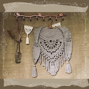 Può includere: Uno scialle di uncinetto grigio con frange appeso a un appendiabiti in metallo. Lo scialle ha un design circolare al centro ed è realizzato con un punto delicato.