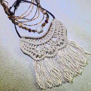 以下が含まれることがあります： クリーム色のかぎ針編みのネックレス。下部にフリンジがあり、茶色とタン色のビーズネックレスが複数連なっています。ネックレスには中央にペンダントがあり、明るい色の表面に表示されています。