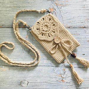 May include: A beige crochet crossbody bag with a decorative floral design on the flap. The bag features a wooden button closure, a tassel, and a long strap with wooden beads. The bag is made of a natural fiber.