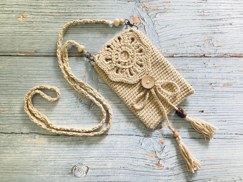 May include: A beige crochet crossbody bag with a decorative floral design on the flap. The bag features a wooden button closure, a tassel, and a long strap with wooden beads. The bag is made of a natural fiber.