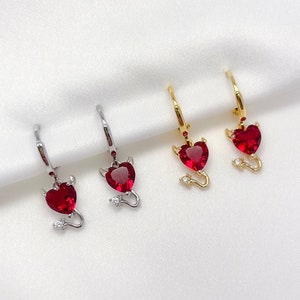 May include: Four pairs of earrings with a red heart and a devil tail charm. The earrings are available in silver and gold.
