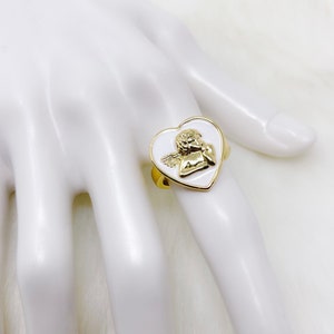 Puede incluir: Anillo de tono dorado con un dise&ntilde;o en forma de coraz&oacute;n blanco que presenta un querub&iacute;n con alas.