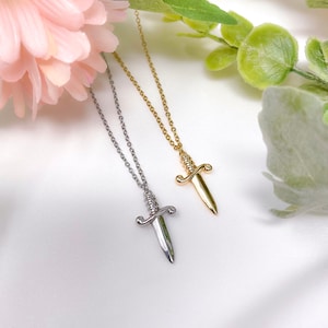 May include: Two silver and gold necklaces with dagger pendants. The daggers have a twisted design and are on a white background.