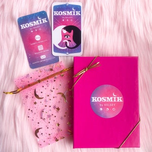 Op de afbeelding: Een roze cadeaudoos met een gouden lint en een witte sticker met de tekst "KOSMIK by KELSEY" en een halve maan ontwerp. De doos staat op een roze pluche oppervlak naast een roze organza tas met gouden sterren en manen. Er zijn twee visitekaartjes met hetzelfde ontwerp en dezelfde tekst als de sticker op de doos.