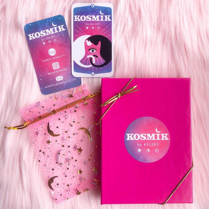 Op de afbeelding: Een roze cadeaudoos met een gouden lint en een witte sticker met de tekst "KOSMIK by KELSEY" en een halve maan ontwerp. De doos staat op een roze pluche oppervlak naast een roze organza tas met gouden sterren en manen. Er zijn twee visitekaartjes met hetzelfde ontwerp en dezelfde tekst als de sticker op de doos.