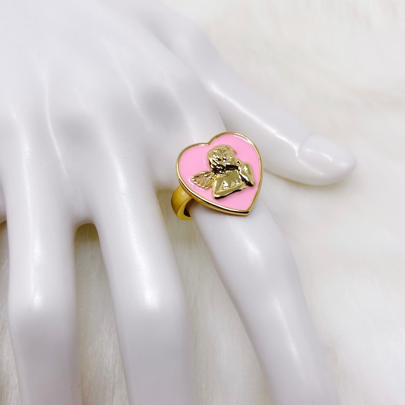 Puede incluir: Un anillo de oro con un dise&ntilde;o en forma de coraz&oacute;n rosa con un querub&iacute;n con alas. El anillo est&aacute; en una mano de maniqu&iacute; blanca.