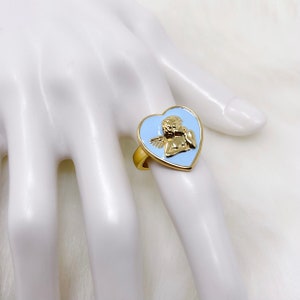 Puede incluir: Un anillo de tono dorado con un engaste en forma de coraz&oacute;n de esmalte azul con un dise&ntilde;o de querub&iacute;n.