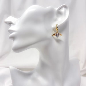 May include: Gold hoop earrings with a dangling heart charm featuring a clear crystal and small gold wings.