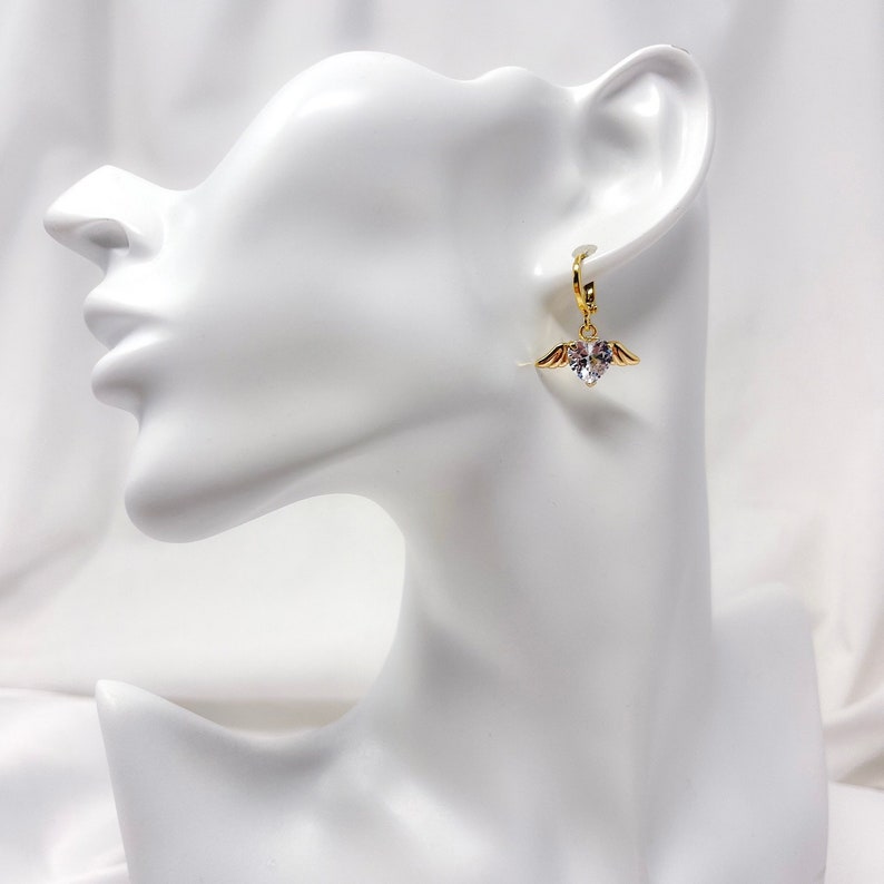 May include: Gold hoop earrings with a dangling heart charm featuring a clear crystal and small gold wings.