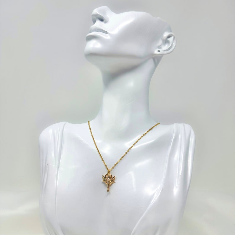 May include: A gold chain necklace with a pendant featuring a stylized key with wings. The pendant is on a white mannequin.