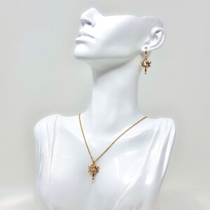 Op de afbeelding: Een gouden ketting- en oorstekerset met een delicaat sleutelhangertje met ingewikkelde details. Het sleutelhangertje heeft een kleine, heldere steen aan de bovenkant.