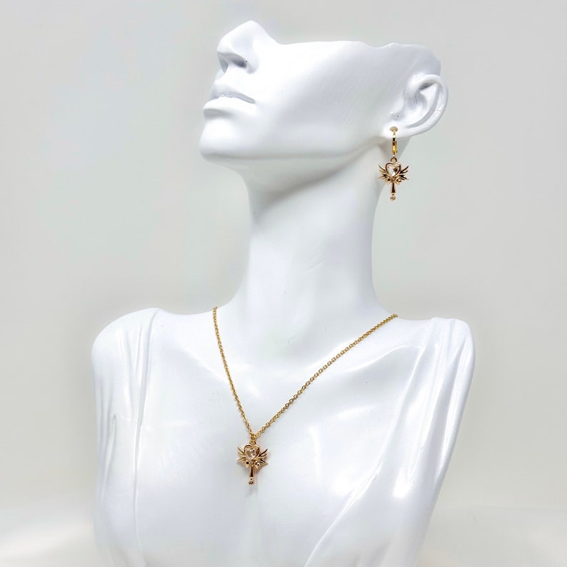 Op de afbeelding: Een gouden ketting- en oorstekerset met een delicaat sleutelhangertje met ingewikkelde details. Het sleutelhangertje heeft een kleine, heldere steen aan de bovenkant.