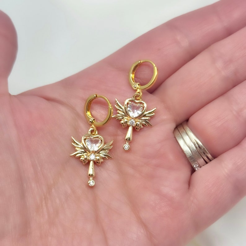 Op de afbeelding: Gouden creolen met een hangende bedel in de vorm van een hartvormig kristal met vleugels. De bedel is versierd met kleinere kristallen.