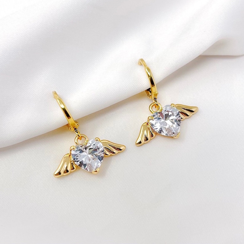 May include: Gold hoop earrings with a dangling heart-shaped crystal accented with gold wings.