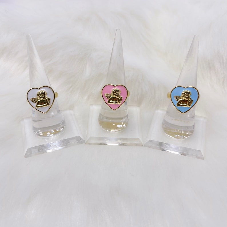 Puede incluir: Tres anillos de oro con engastes en forma de coraz&oacute;n que presentan un dise&ntilde;o de querub&iacute;n. Los anillos se muestran en soportes para anillos de pl&aacute;stico transparente. Los corazones son blancos, rosas y azules.