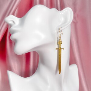 May include: Gold sword earrings with a detailed design. The earrings are hanging from a mannequin's ear.
