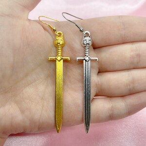 May include: A pair of sword earrings with a lion head design. One earring is gold and the other is silver.