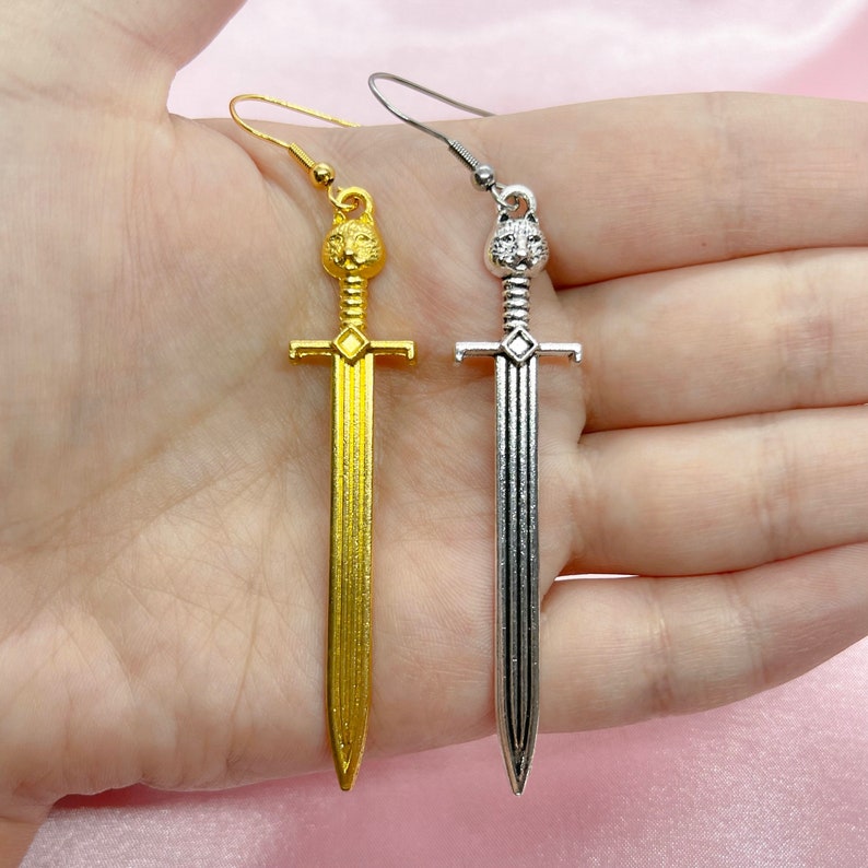 May include: A pair of sword earrings with a lion head design. One earring is gold and the other is silver.