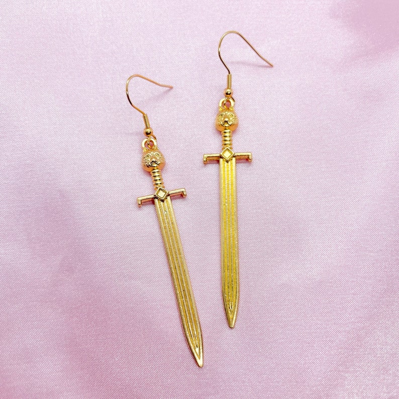 May include: A pair of gold sword earrings. The swords have a detailed design and a small crown at the top.
