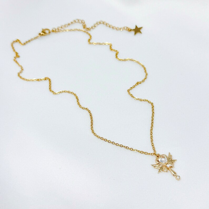 May include: A gold chain necklace with a pendant shaped like a wand with wings and a clear crystal in the center.