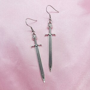 May include: A pair of silver sword earrings with a detailed design. The swords have a pointed tip and a decorative handle.