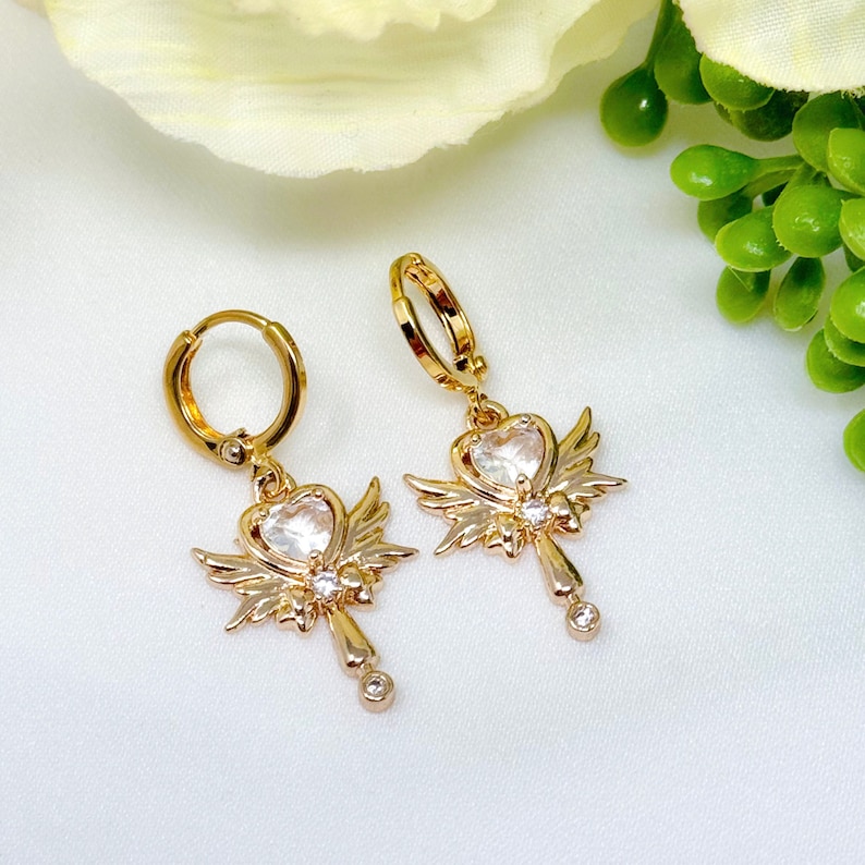 Op de afbeelding: Een paar gouden creolen met een hangende hartbedel versierd met heldere strass steentjes en kleine vleugels. De hartbedel heeft een kristal-achtige middelste steen.