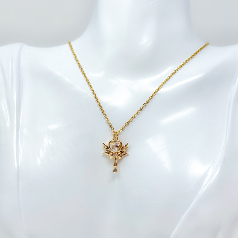May include: A gold necklace with a heart-shaped pendant featuring a clear crystal and angel wings.